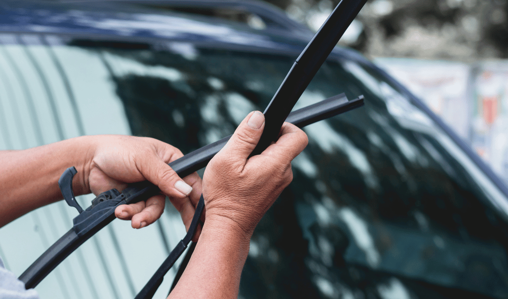 A person replaces a car windshield wiper, holding the new wiper blade in one hand and the arm mechanism in the other.
