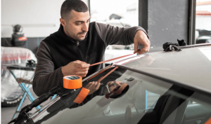 A technician applies tape along the edge of a car's windshield in a garage, focusing on auto glass repair